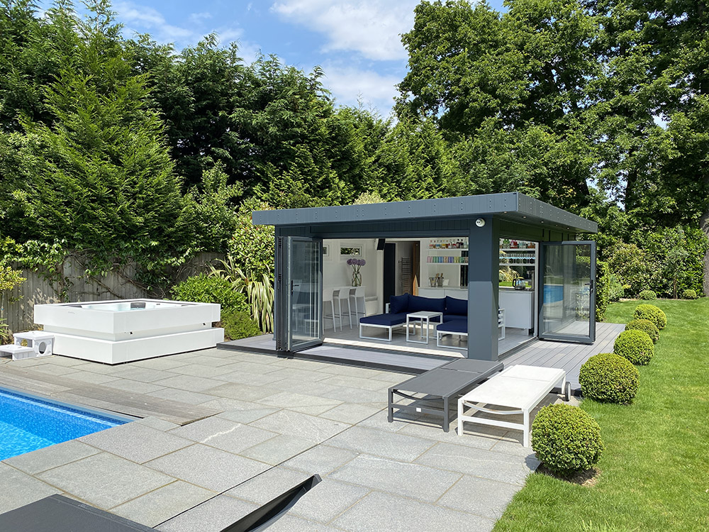Poolside garden building sitting next to a hot tub and swimming pool. Inside the poolside room, you can see deck chairs, a bespoke bar and a skylight in the roof.